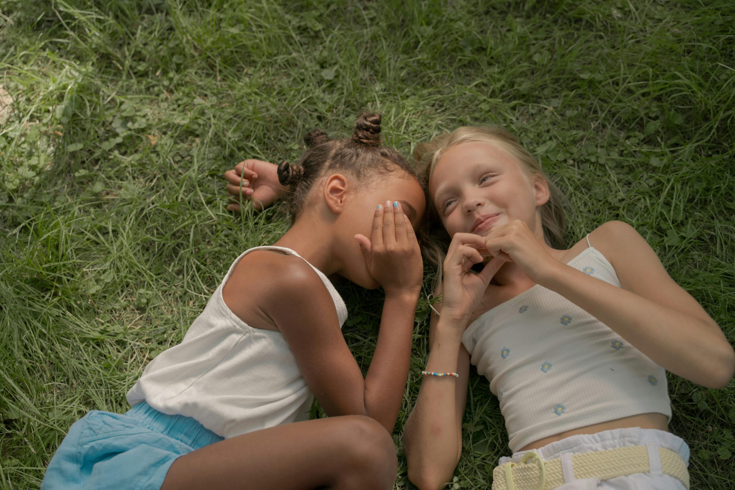 Two teenagers lying on grass whispering and smiling in a summer setting.