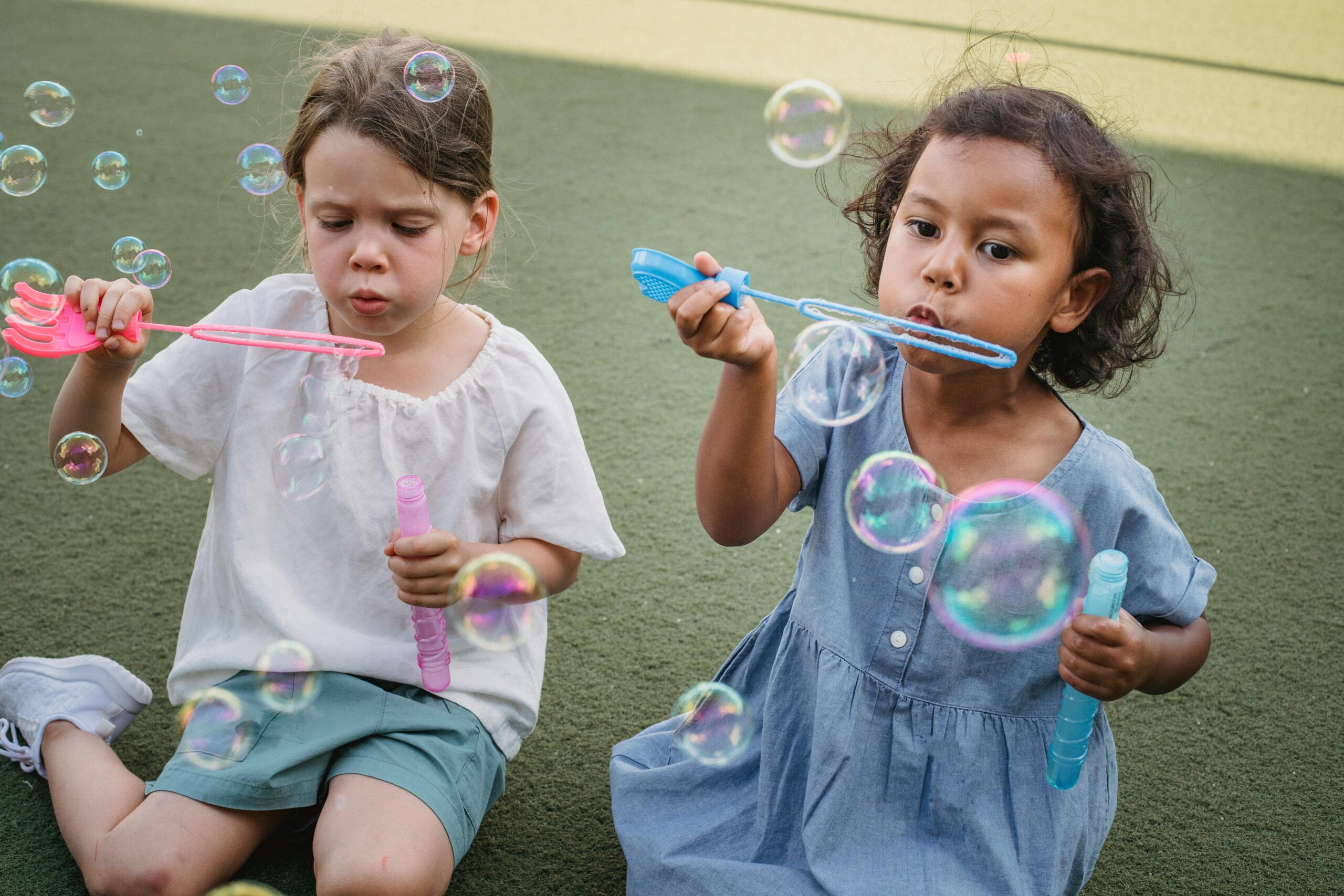 Two young girls enjoy a playful summer day blowing bubbles outdoors on a grassy field.